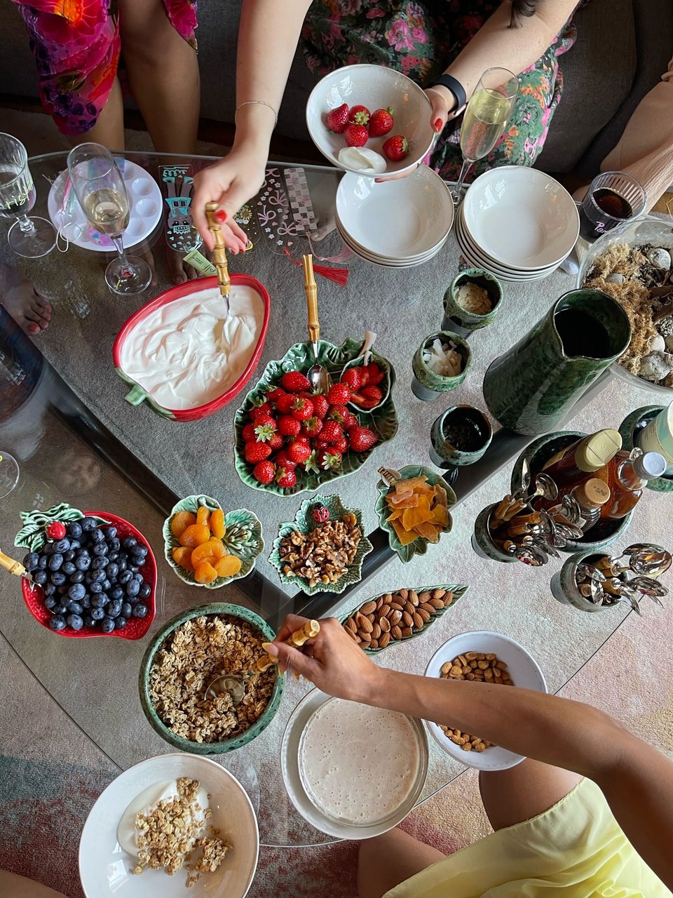 A coffee table covered in bowls of fresh fruit, granola, yogurt, nuts, and drinks, with people serving themselves.