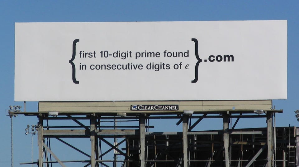 A photograph of a billboard. There is no image in the advertisement, just the following text: {first 10-digit prime found in consecutive digits of e}.com