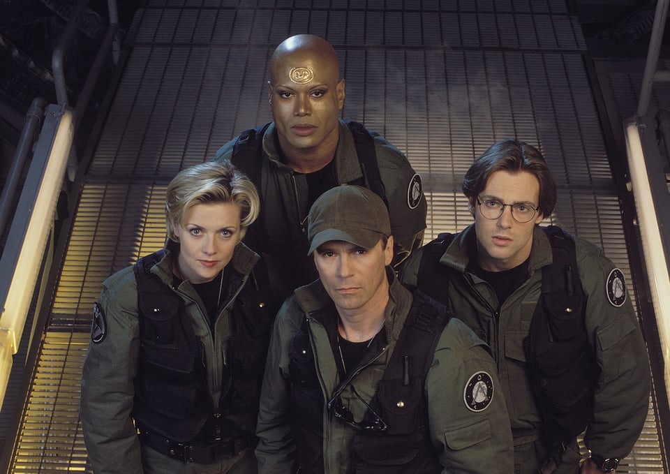 Image of the four starting main characters of Stargate: SG-1, all wearing military uniforms.  One is a blonde woman with short hair; one is a white man wearing a hat; one is another white man with floppy brown hair, glasses, and a face that’s just begging to die upwards of ten times over the show’s run; and one is a bald Black man with heavy eyeshadow and a golden symbol on his head.