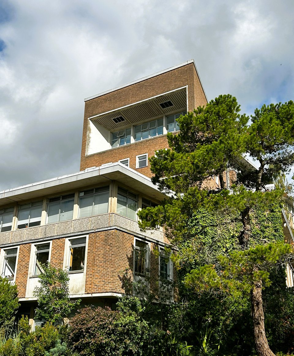 Two 1960s brick and concrete buildings. In the foreground, half hidden by trees, is a two storey block set on stilts with a mix of brick and concrete walls. Behind it is a brick building with two massive double-height balconies deeply set into them.