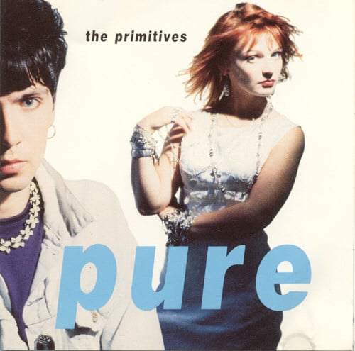 A color photograph of two members of The Primitives, both looking at the camera. One is a white man with short black hair, an earring, and necklace made of metal butterfly shapes. He is close to the camera and the left side of his face is cropped off. To the right and behind him is a white woman with red hair in a white dress. Her left arm is wrapped around her waist and her right arm is lifted so she's touching her right shoulder with her right hand. The background is white.