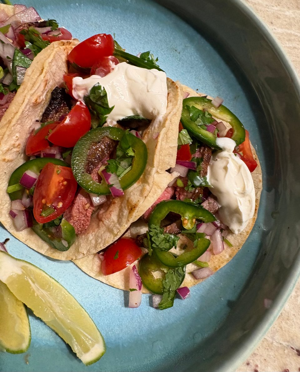 Tandoori steak tacos loaded with fresh tomato, red onion, jalapeño, cilantro, and limes on a blue plate.