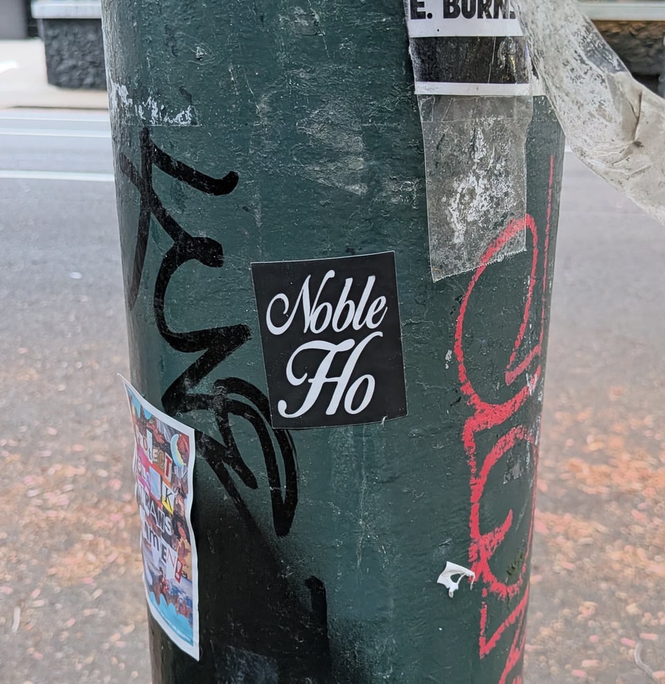 A close up photo of a green lamppost with a square black sticker on it. The sticker says NOBLE HO in flourished script.
