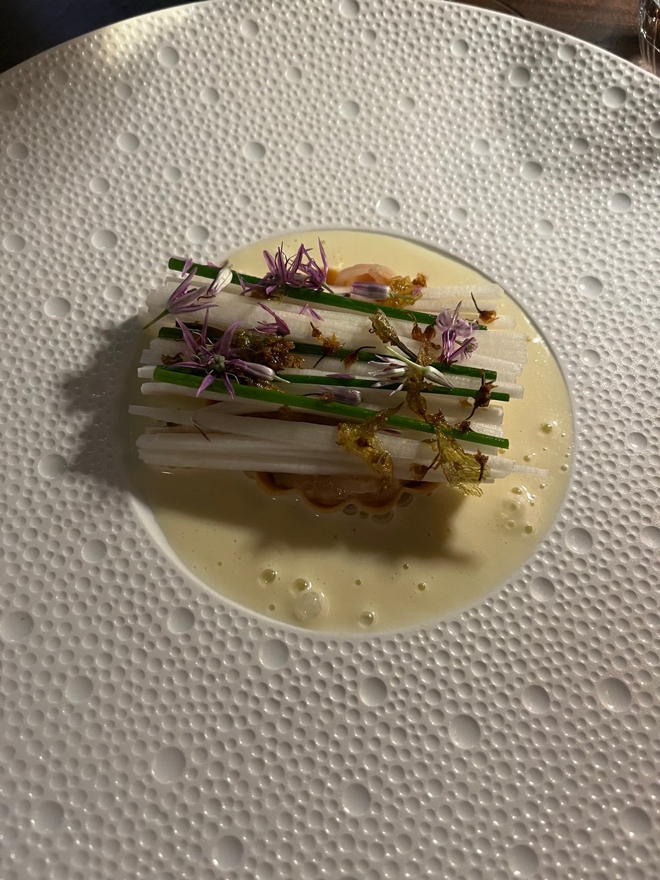 Julienned vegetables with edible flowers sitting in a creamy white sauce, in a dish that is decorated with white bubbles