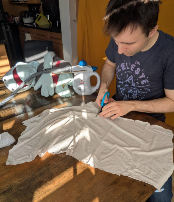 Kurt cuts an old t-shirt into a cloud shape. Behind him, a steamer is visible. Beside that, Herman the puppet is flopped dramatically on the table like a Victorian woman on a fainting couch.