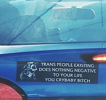 Photo of a bumper sticker that is black with white text that reads “trans people existing does nothing negative to your life you crybaby bitch”