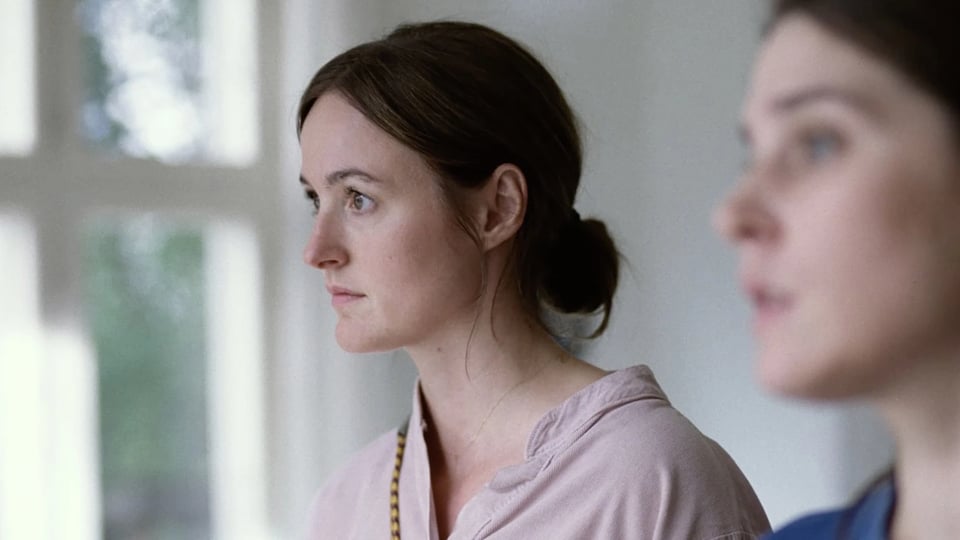 A woman with dark brown hair tied back in a neat bun, wearing a light lavender shirt, staring to the left. The background is blurred out, as is another woman in the foreground.