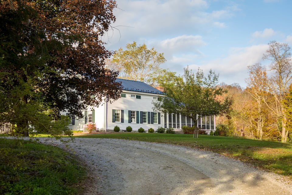 A two-story farmhouse sits beyond a curved dirt driveway, partially obscured by trees. It has white siding with green shudders and a gray metal roof.