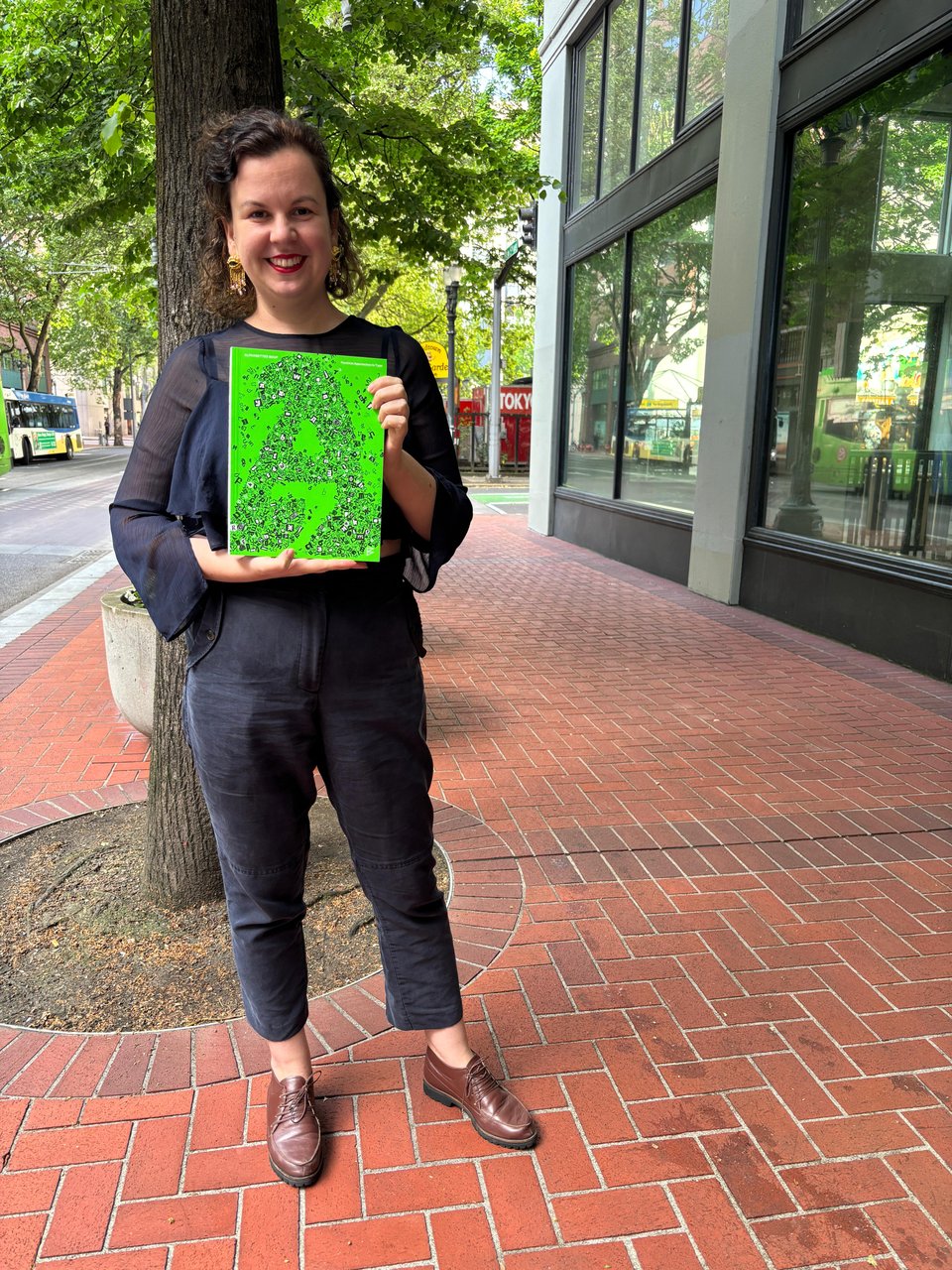 a person holds a green book smiling on a paved brick sidewalk on a city street next to a tree