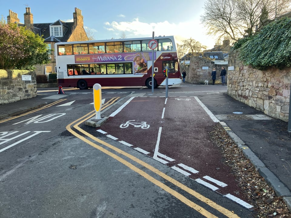 The mouth of a side road is pictured meeting a main road where a red and white double-decker bus is passing by; two people and a dog on the other side of the street wait to cross towards us at a newly installed toucan crossing. In front of us, a red asphalt bi-directional cycle lane stretches for a few metres; it is separated from the side road's carriageway by a kerb and light bollard. Markings on the side road and a sign on a post indicate there is no right turn for motor vehicles exiting this road.