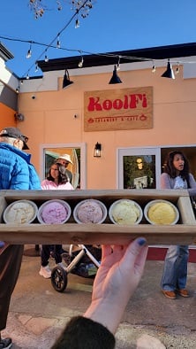 Photo of Nicole’s hand holding up a tray of five scoops of ice cream in front of a doorway and sign to KoolFi Creamery.