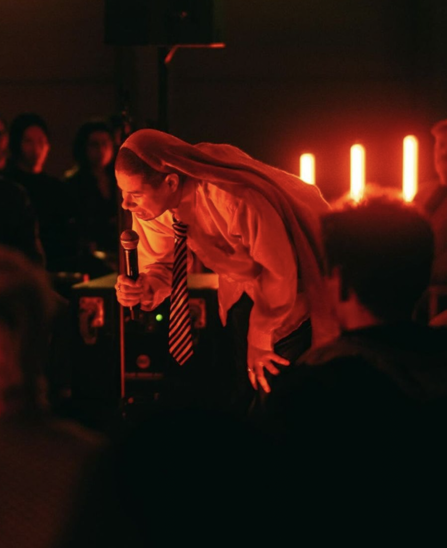 ãssia ghendir, bathed in orange light, wearing leather trousers, school uniform shirt and tie, and a head covering, leans over, eyes closed, while singing into the microphone