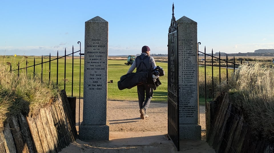 Simon walking through Royal West Norfolk war memorial gates from the beach, decorative wrought iron supported by two stone pillars, links and salt marshes ahead
