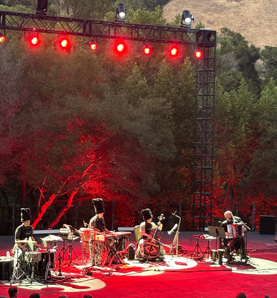 Alt text: Four people, three in big hats, play instruments in front of trees and hills under red lights.