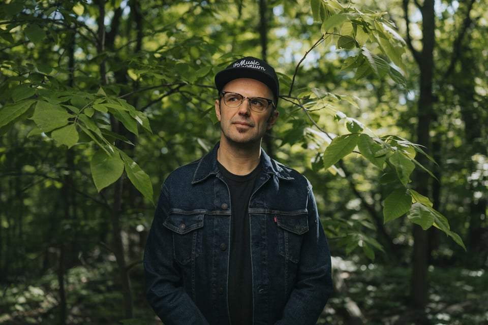 A man with glasses stands amid trees. He's wearing a dark blue denim jacket and a black baseball cap.