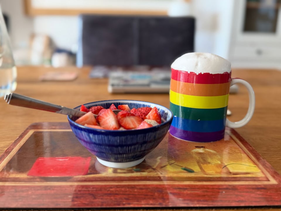A bowl of overnight oats covered in cut up strawberries sits next to a mug filled with coffee and frothy milk on a placemat on a dining room table.