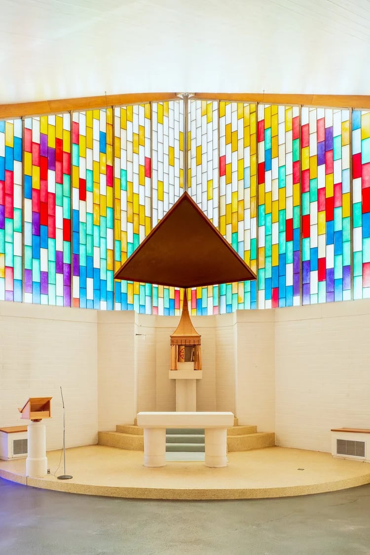 A light-filled church interior where the simple altar is in the corner. The single storey walls are plain, and above them rises windows made up of coloured blocks that form a stylized sun.