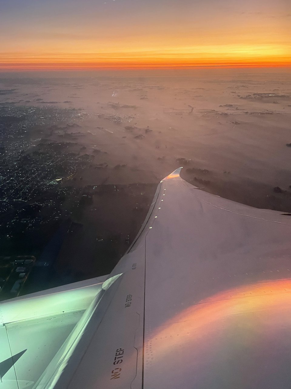 The morning sunrise approaches the horizon. The airplane wing stretches out over the countryside visible under the wing. The countryside is covered in ground fog.