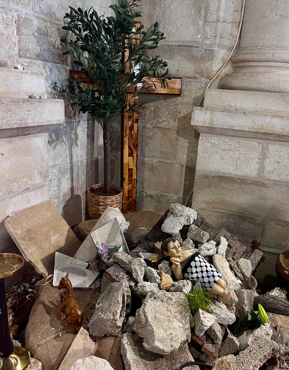 A display of rubble, with a baby (Jesus) lying atop the rubble, covered with a keffiyeh. A large cross and stone walls