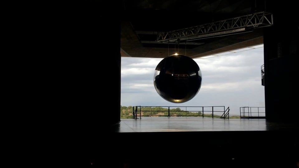 A dark stage with a round glass-like ball hanging over it. The ball represents the first nuclear bomb created by Oppenheimer's team in WW2. Behind the stage, it's open to the outside and the Jemez Mountains are in view. Clouds cover the sky.