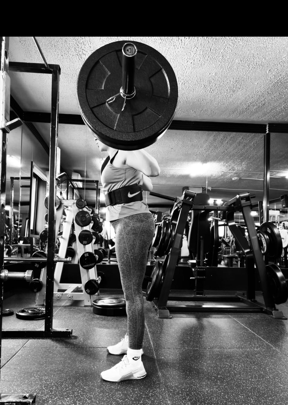 black and white image of a woman seen from the side, standing up and holding very big barbells on her shoulders 
