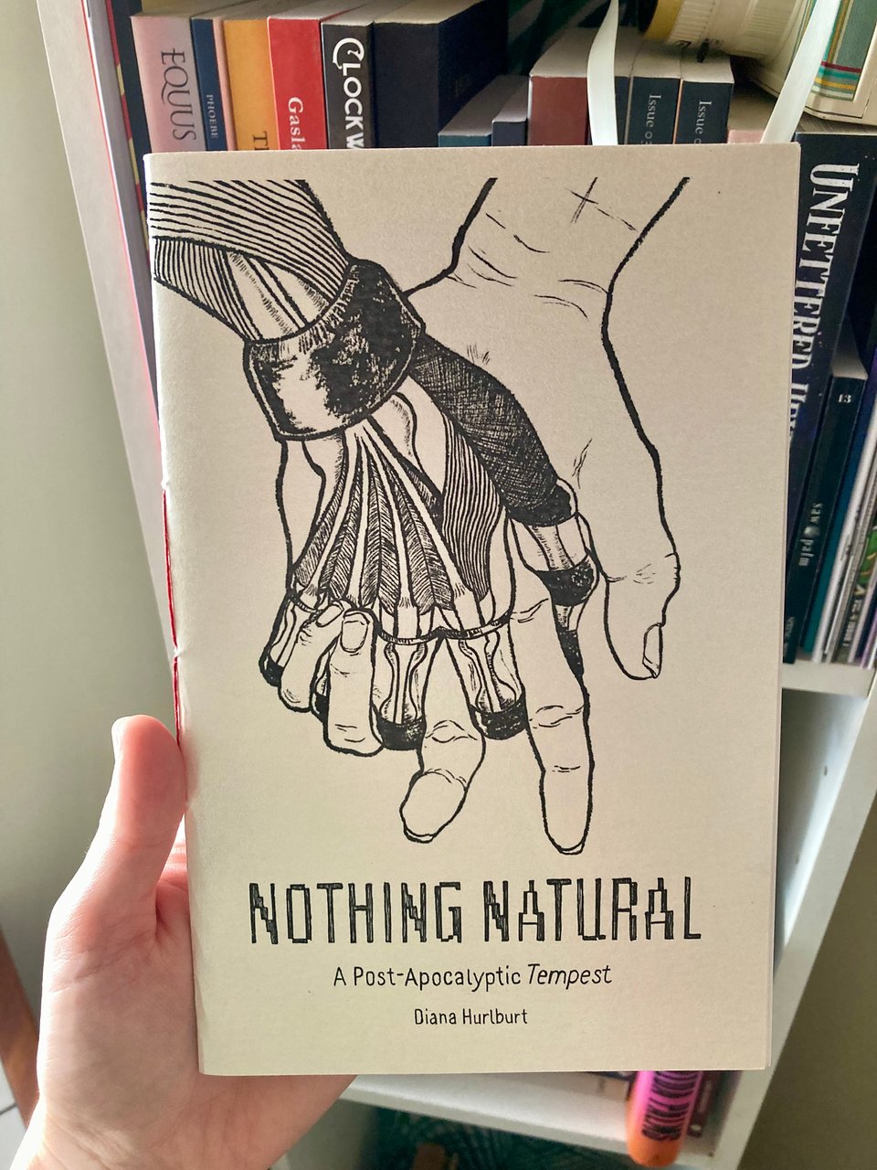 a hand holding the mini-chap of Nothing Natural in front of a bookshelf. the chap is pearlescent white cardboard with a black ink drawing of two hands intertwined: a human and a robot. it's titled "Nothing Natural: A Post-Apocalyptic Tempest."