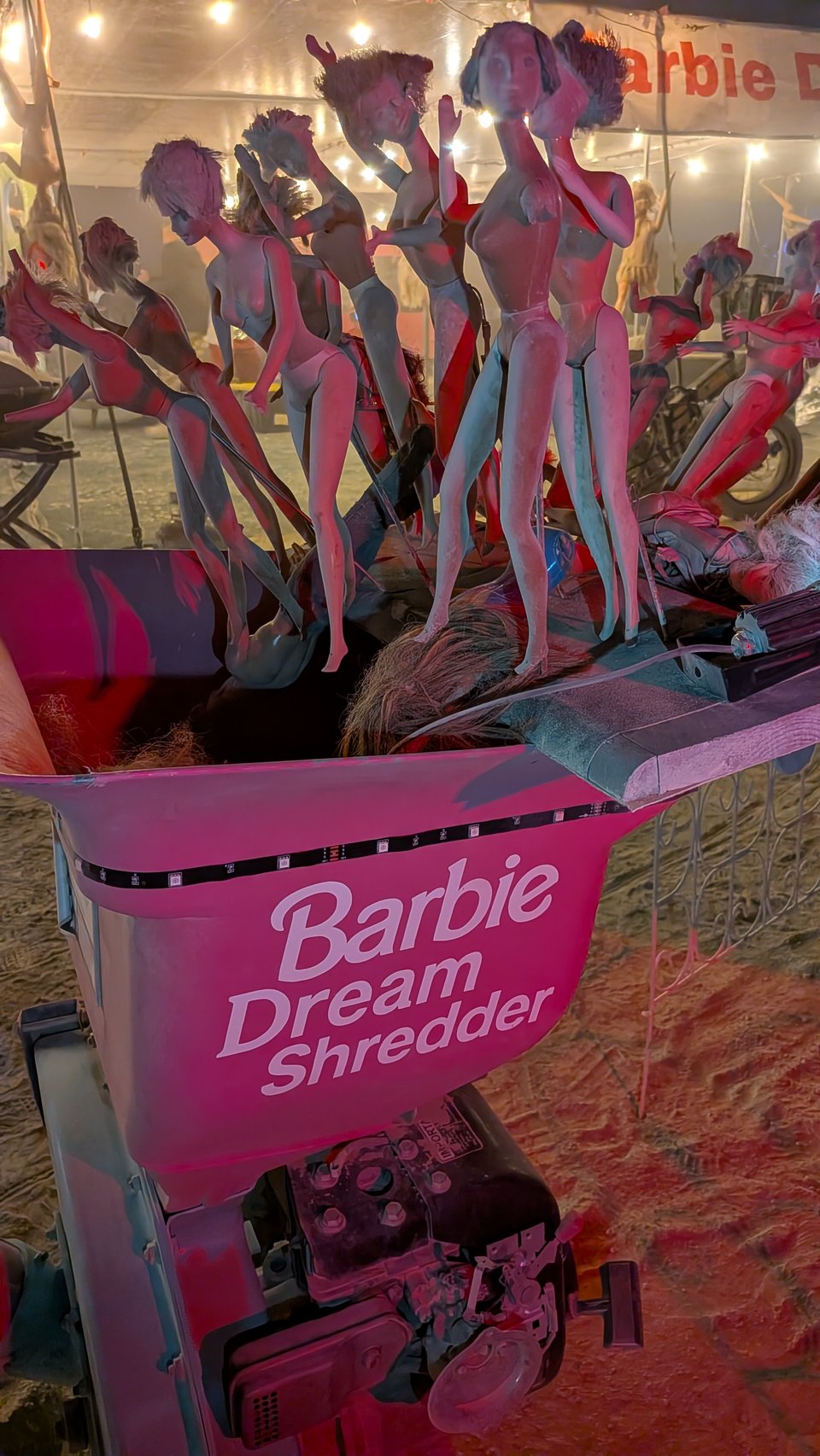 A motionless conveyer belt covered in naked dolls being fed into a machine labeled "Barbie Dream Shredder" in the classic pink and white Barbie colors.