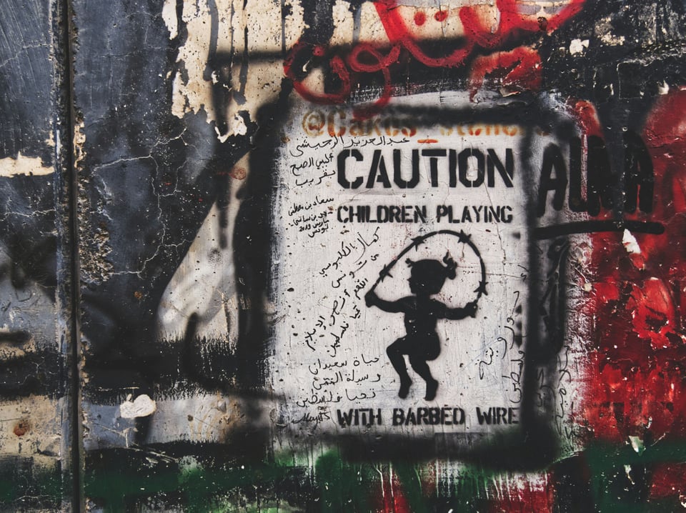 Graffiti on the apartheid wall in Palestine painted in black on white background. It shows a child jumping rope with barbed wire and says "CAUTION: Children playing" above the girl jumping rope and "with barbed wire" below it.