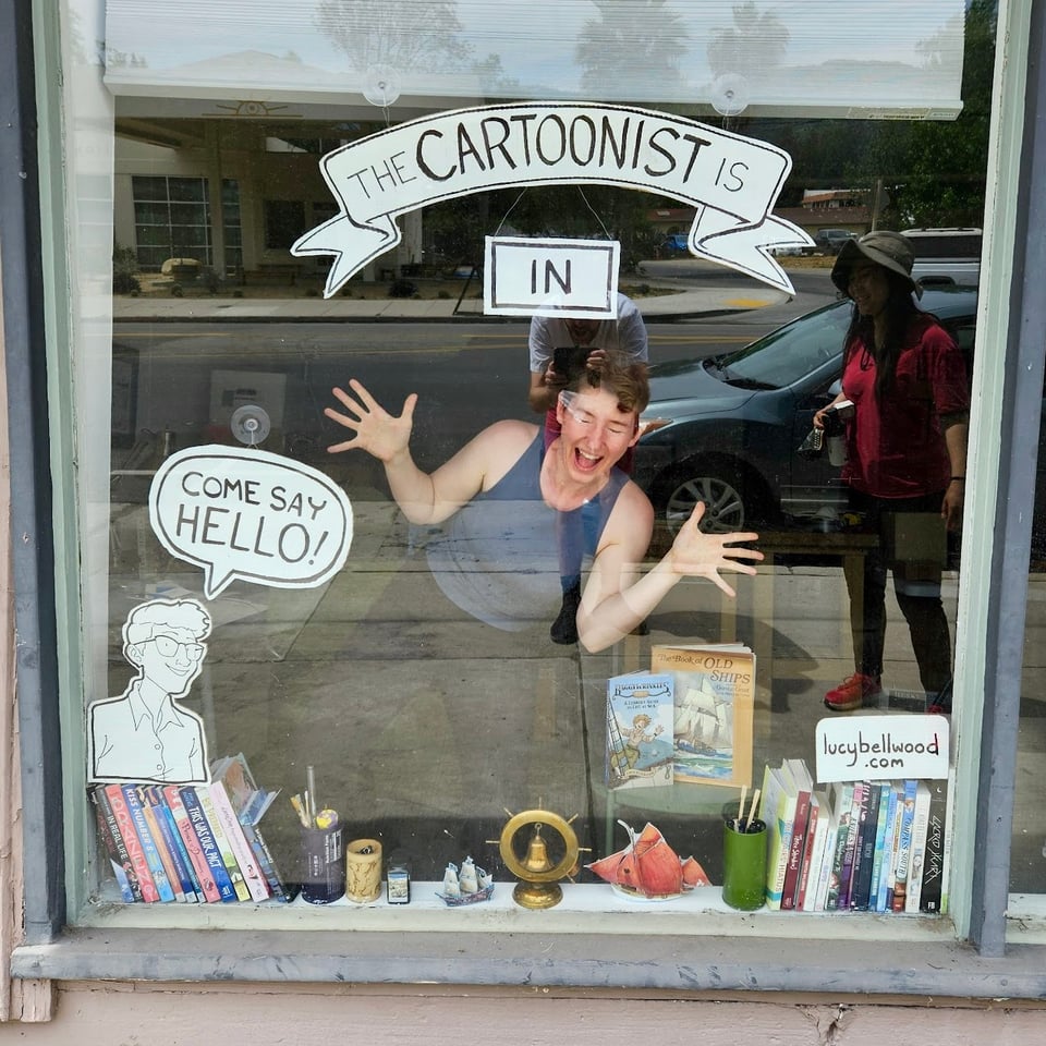 Lucy beaming in the window of her studio under a sign saying "The cartoonist is in!"