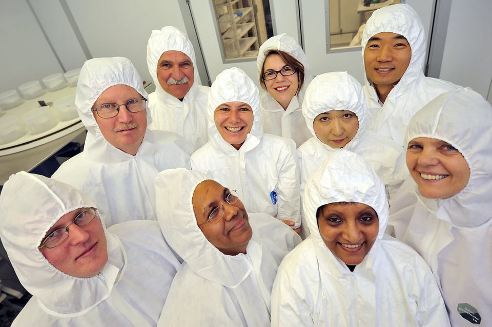 A group of 10 smiling lab staff members, dressed in white clean room garb