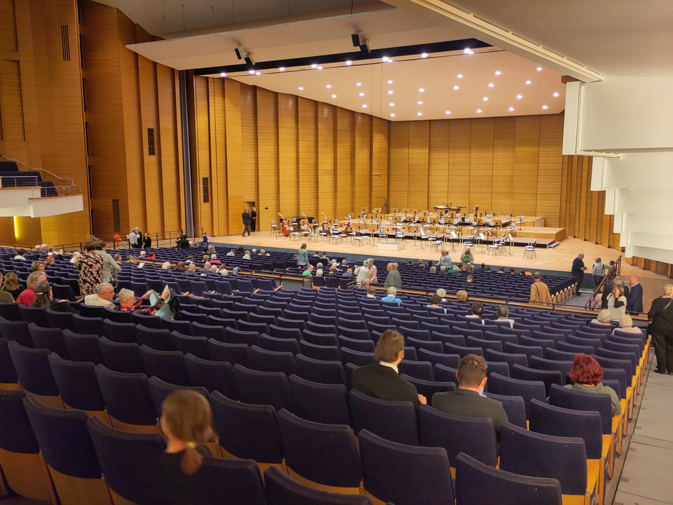 Blick in einen Konzertsaal. vereinzelt sitzen Menschen in den langen Sitzreihen, auf der Bühne stehen die Stühle eines Sinfonieorchesters.