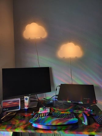 A desk with a laptop and second monitor. On the wall behind it are two lit cloud-shaped lights. The entire area is covered in rainbows from prisms on the windows out of the frame.