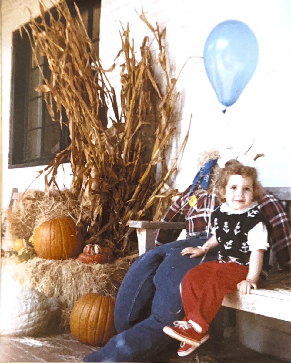 A young girl sits on a bench next to an apparently headless scarecrow