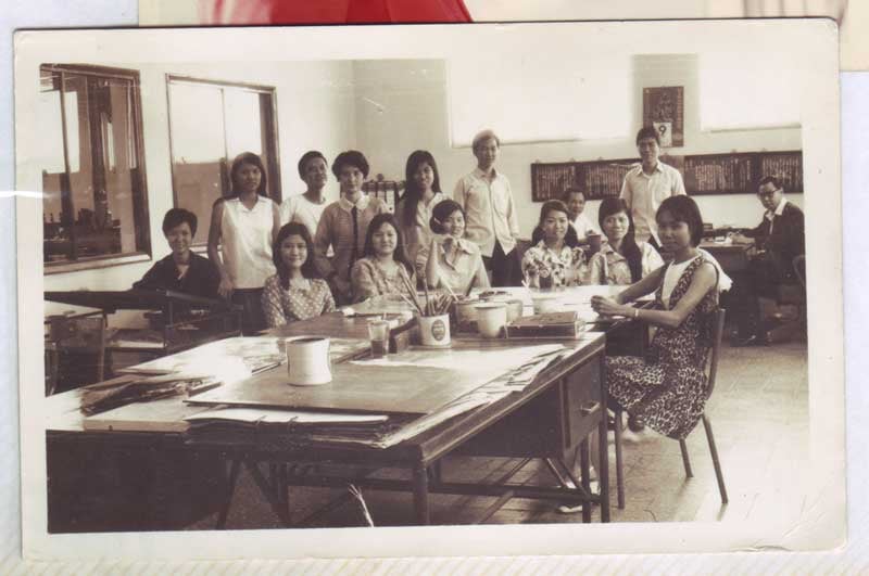 a sepia toned photo of young viet adults in their twenties posing behind a large classroom table. daily calendar in the background.