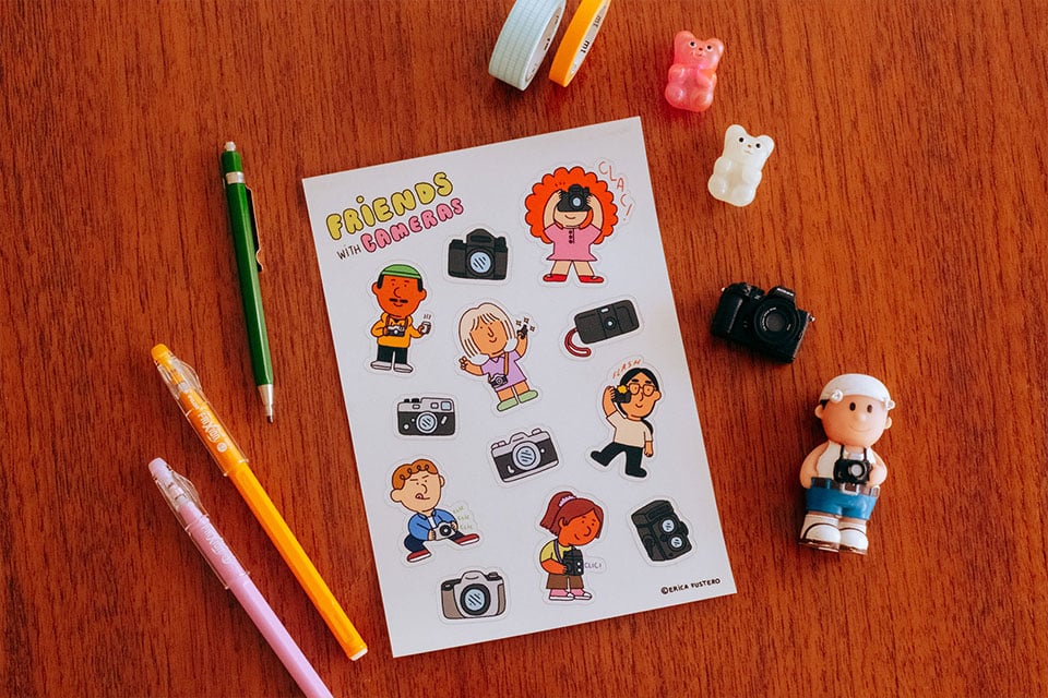 Fotografía de una hoja de pegatinas sobre una mesa, rodeada de material de papelería y miniaturas.
Photograph of a sheet of stickers on a table, surrounded by stationery and miniatures.