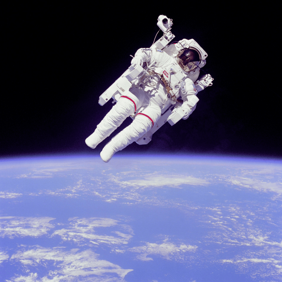 A photo of an astronaut floating untethered in space, wearing a white spacesuit that appears to have some sort of mechanism behind it for propelling itself around through space. Behind the astronaut is a completely black area of space and below it the bright blue glow of earth, covered in a small amount of thin white clouds.