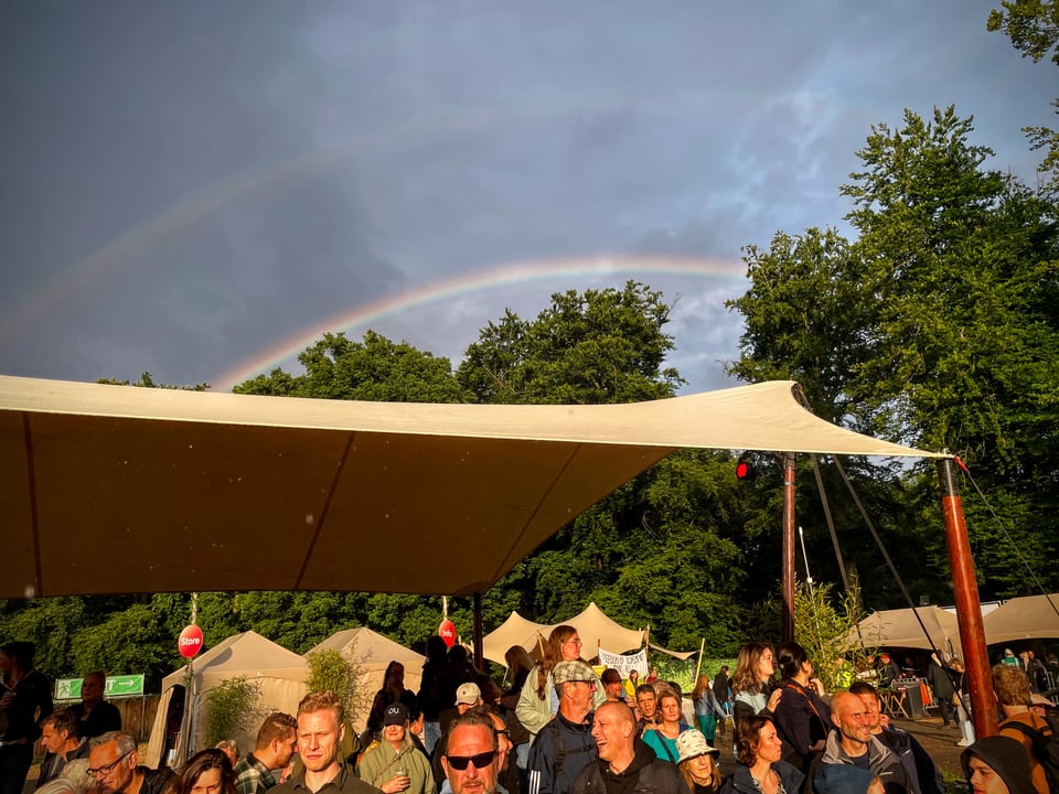 Two rainbows stretch over the background behind a row of trees. In front there is a high tented area protecting some of the crowd from rain.