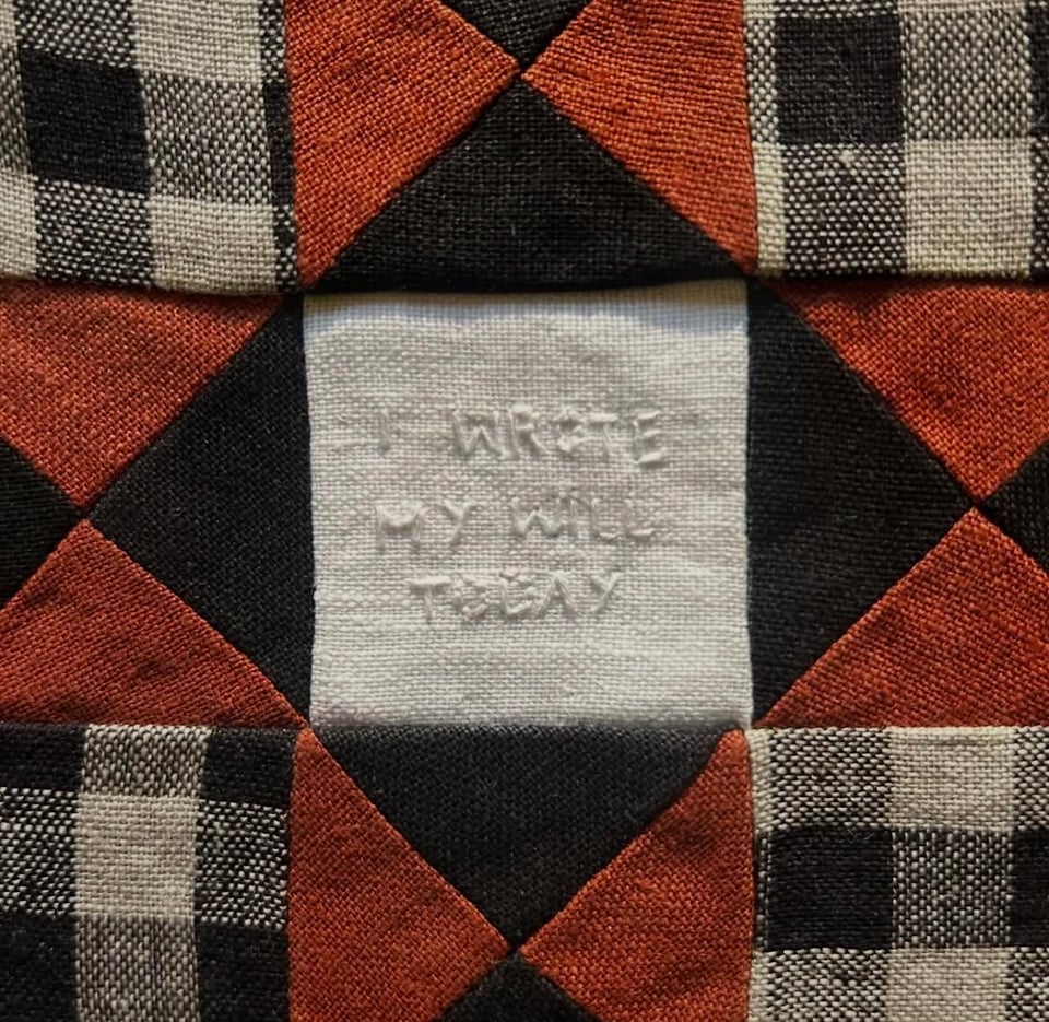 A black, red, and white quilt of geometric patchwork. "I wrote my will today" is stitched into the center white square with white thread.