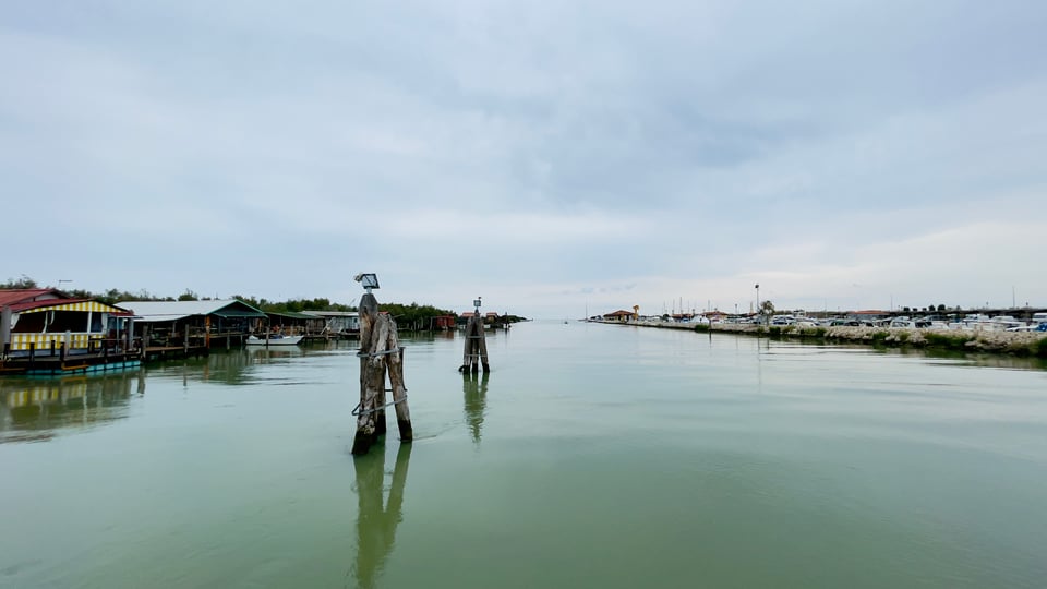 A branch of the Po river delta, looking out to sea. Gray sky above, green water in the foreground. Fishing huts on stilts on the left bank, parked cars on the right bank. Sailboats from the local marina can be spotted farther away along the right bank