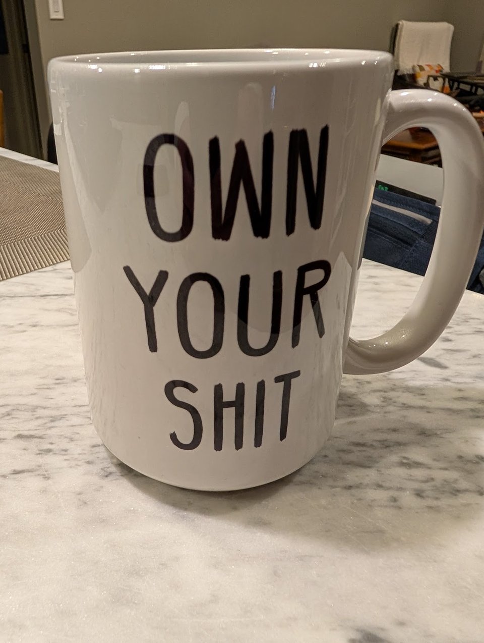 A white coffee mug sitting on a white marble counter. The words "own your shit" are written on the mug.