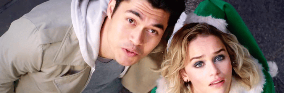 A still from Last Christmas. A white woman dressed as an elf and a very handsome Asian man in a hoodie stare upwards.