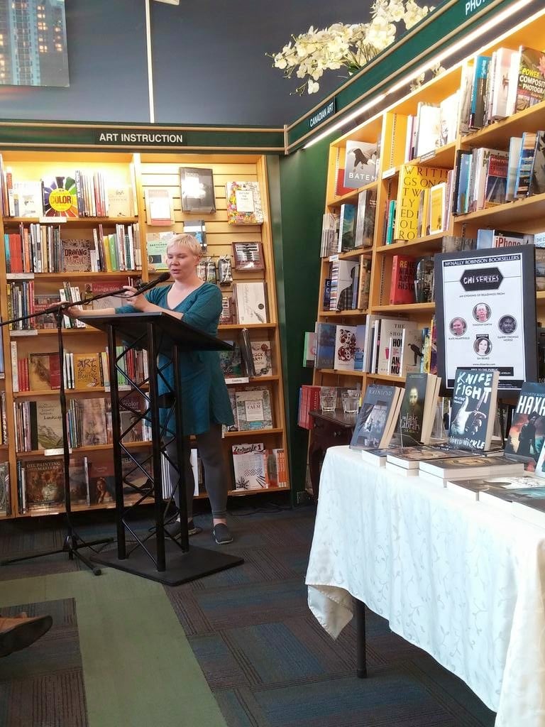 Kate reading in a blue dress with short blonde hair, in a corner lined with bookshelves.