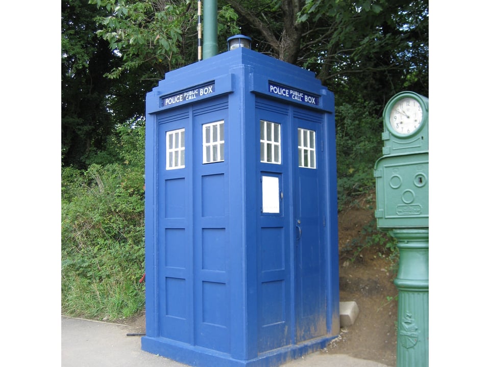 A large, square, bright blue box labeled "POLICE PUBLIC CALL BOX" at the side of a road. Next to it is some kind of gauge.