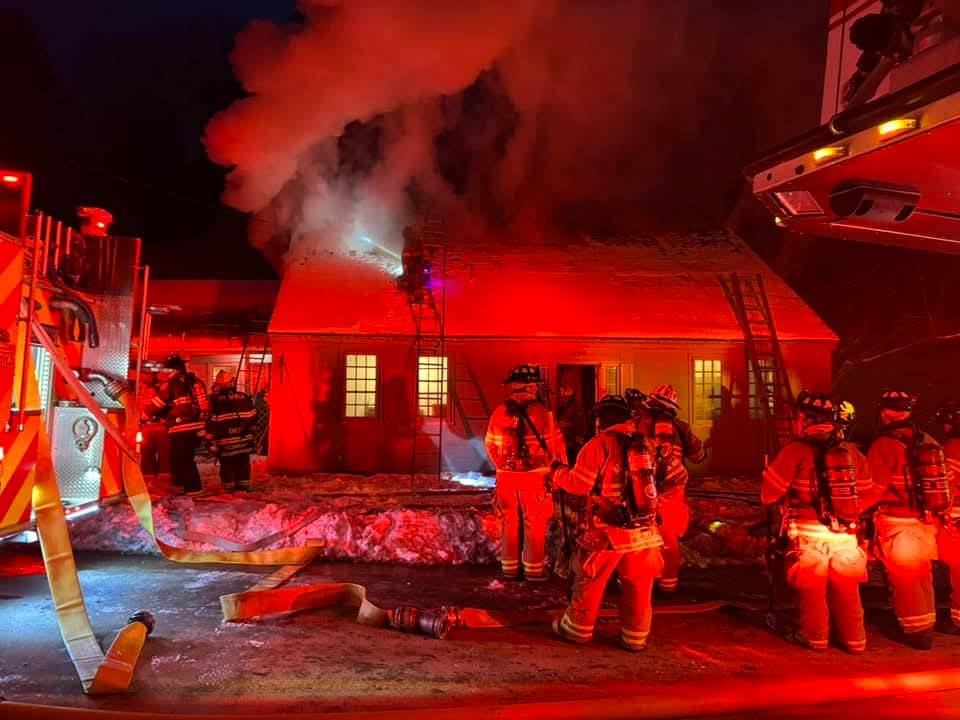 An emergency scene at night with firefighters responding to a structure fire. There is visible smoke rising from the building, indicating active flames inside. Fire trucks are parked nearby, and personnel in full protective gear are working on extinguishing the fire.