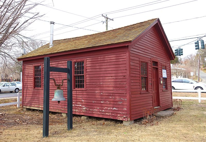 A picture of a one-room schoolhouse painted red in Simsbury, CT on the Massacoh Plantation. It is a small, squat building painted red, with two windows on one side and a window and a doorway in the front. The tile roof looks golden. It's in the middle of a grassy empty field with cars going by. There is a big school bell on a frame. Photo by Daderot/Wikimedia