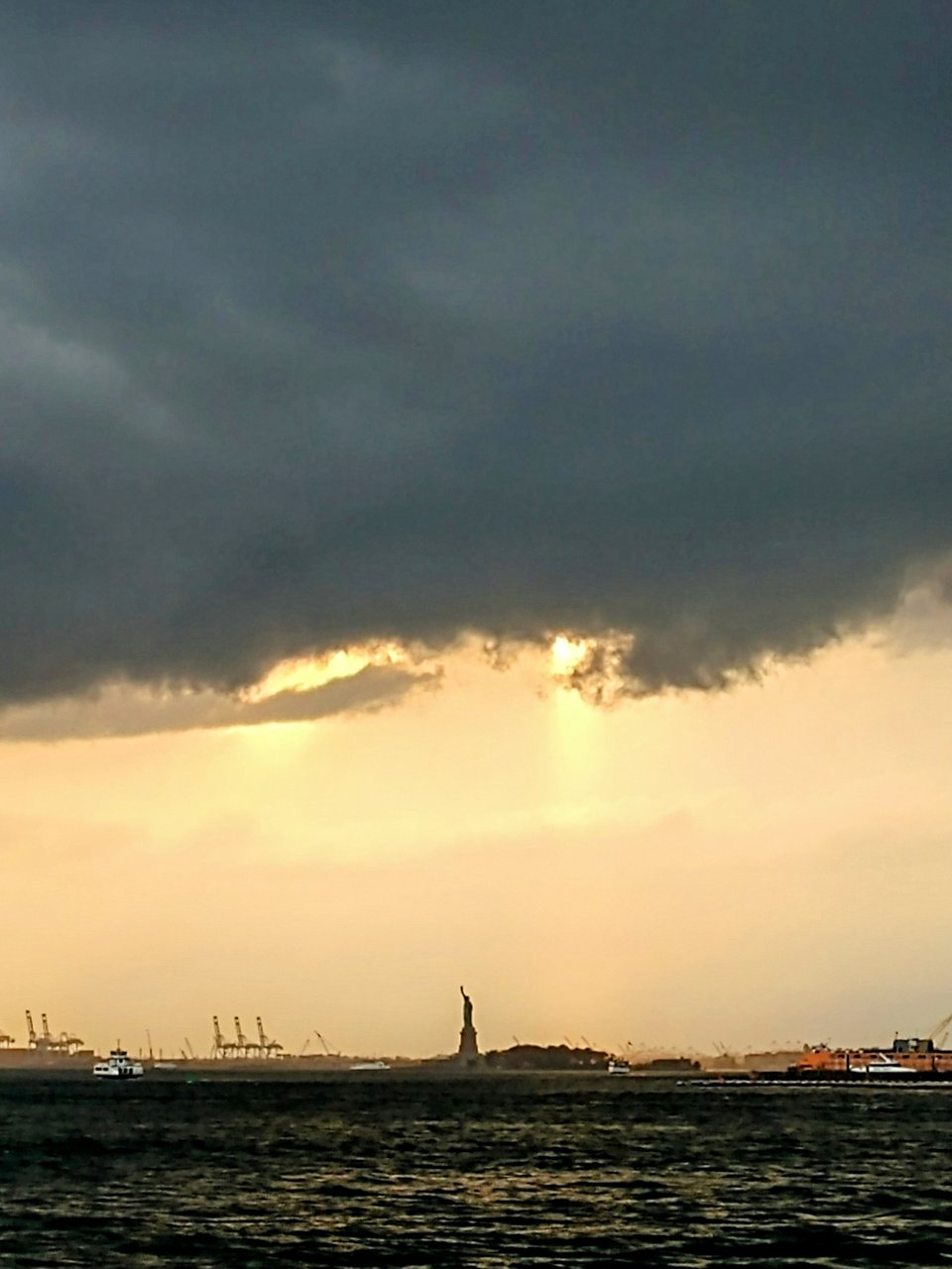 The Statue of Liberty under a beam of yellow sunlight breaking through charcoal colored storm clouds