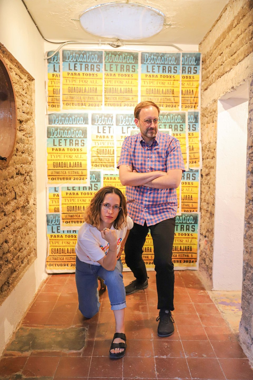 Zrinka is kneeling with a fist raised in front of Jesse, arms crossed and leaning forward with a grimace. Background is a wall of lucha style posters that say "Letrástica".