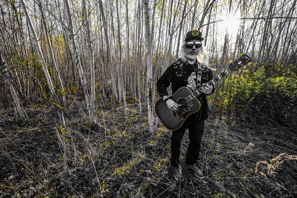 Singer J Mascis stands among trees holding an acoustic guitar and wearing mirrored sunglasses and a baseball cap