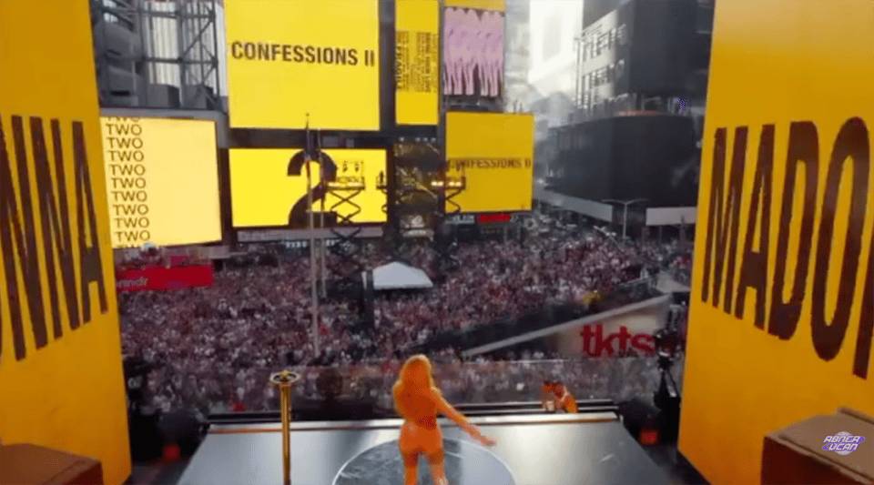 Madonna stands on a small stage elevated above Times Square and looks out over a massive crowd. Various signs around the square say "Confessions II", "Madonna", "Two" and "2".