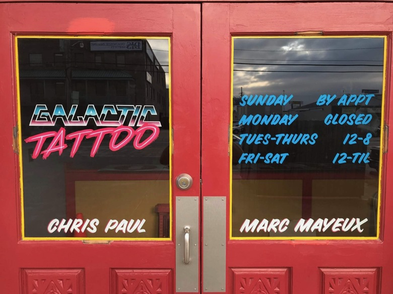 Two hand painted windows of a red double door. On the left is a logo “Galactic Tattoo” in an 80s space and pink neon aesthetic; at the bottom is the name “Chris Paul”. On the right are the open hours and the name "Marc Mayeux"